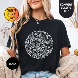 May include: Black t-shirt featuring a white volleyball design with floral and geometric patterns. The shirt has a round neck and short sleeves. The text "Comfort Colors 1717" is visible on the upper right. The text "Ships Next Day" is visible on the upper left.