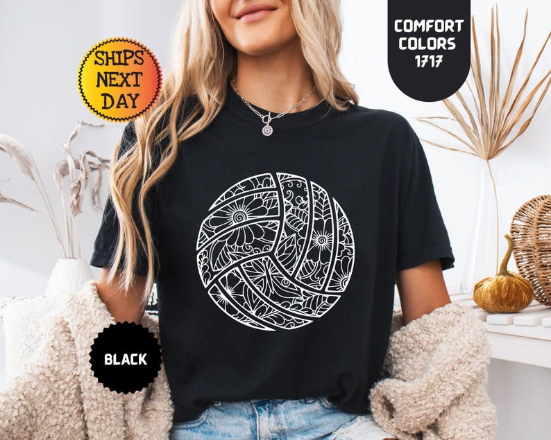 May include: Black t-shirt featuring a white volleyball design with floral and geometric patterns. The shirt has a round neck and short sleeves. The text "Comfort Colors 1717" is visible on the upper right. The text "Ships Next Day" is visible on the upper left.