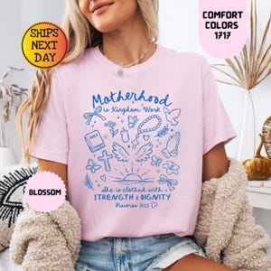 May include: A light pink t-shirt with the words "Motherhood is Kingdom Work" and other religious graphics in blue. The shirt also has the text "She is clothed with Strength & Dignity" and "Proverbs 31:25".