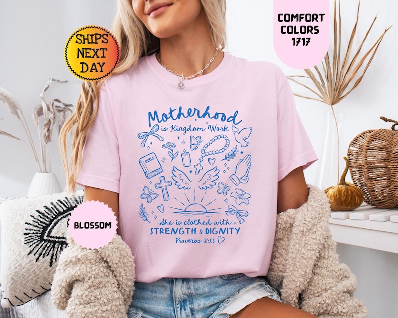 May include: A light pink t-shirt with the words "Motherhood is Kingdom Work" and other religious graphics in blue. The shirt also has the text "She is clothed with Strength & Dignity" and "Proverbs 31:25".
