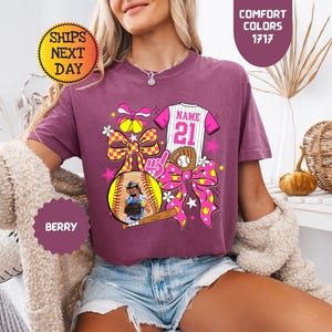 May include: A berry-colored t-shirt featuring a graphic print of a softball, a baseball jersey with the text NAME 21, a baseball glove, a bat, and large yellow and pink patterned bows. The design includes stars and flowers in a vibrant color palette.