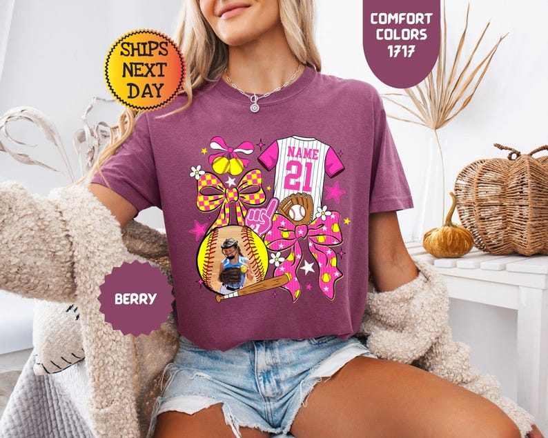 May include: A berry-colored t-shirt featuring a graphic print of a softball, a baseball jersey with the text NAME 21, a baseball glove, a bat, and large yellow and pink patterned bows. The design includes stars and flowers in a vibrant color palette.