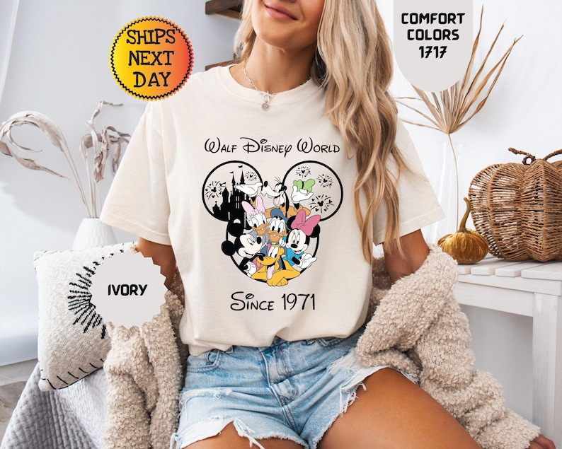 Pode incluir: Camiseta cor marfim com um gr&aacute;fico do Mickey Mouse e amigos, e o texto "Walt Disney World Since 1971". A camisa tem gola redonda e mangas curtas. A camisa tamb&eacute;m tem o texto "Comfort Colors 1717" e "Enviamos no dia seguinte".