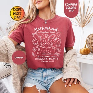May include: Crimson-colored t-shirt with the text "Motherhood is Kingdom Work" and other religious symbols in white. The shirt also has the text "She is clothed with STRENGTH & DIGNITY Proverbs 31:25".
