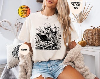 May include: An ivory short-sleeve t-shirt featuring a black ink-style graphic of a skeleton sitting cross-legged in a field of flowers while reading an open book. The shirt has a crew neckline and is made of cotton fabric with a relaxed fit.