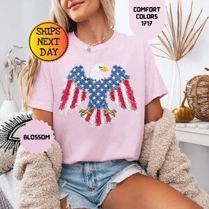 May include: A light pink t-shirt featuring an American flag eagle design. The eagle has blue wings with white stars and red and white striped feathers. The shirt has the text "Comfort Colors 1717" and "Blossom".
