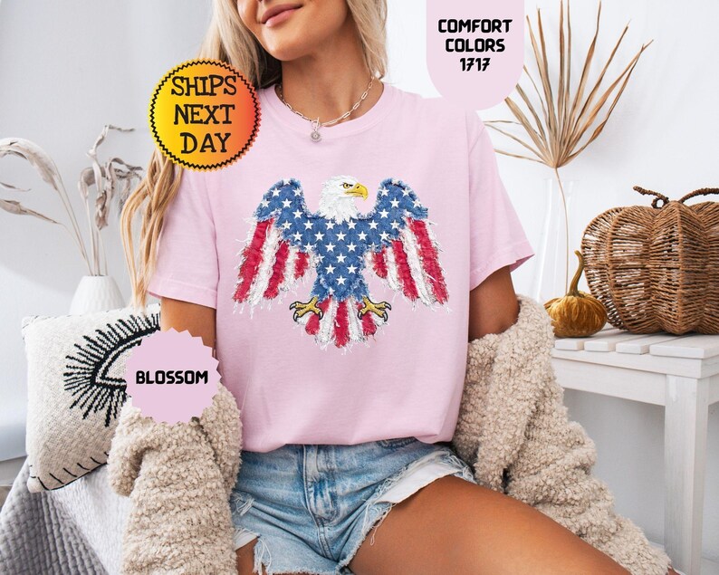 May include: A light pink t-shirt featuring an American flag eagle design. The eagle has blue wings with white stars and red and white striped feathers. The shirt has the text "Comfort Colors 1717" and "Blossom".