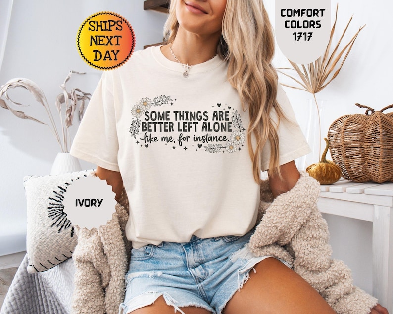 May include: Ivory-colored t-shirt with the text "SOME THINGS ARE BETTER LEFT ALONE like me, for instance" in black script and floral accents. The shirt is part of the Comfort Colors collection.