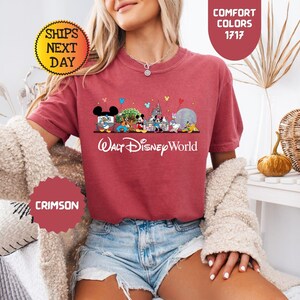 May include: Crimson-colored t-shirt featuring a Walt Disney World graphic. The design includes various Disney characters and landmarks. The text "Walt Disney World" is printed in white script below the graphic. The shirt has short sleeves.