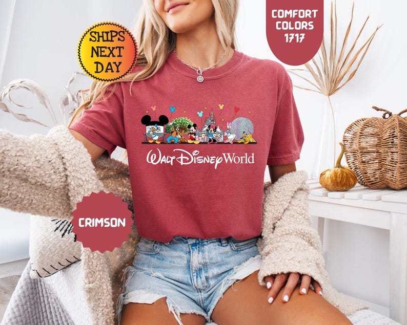 May include: Crimson-colored t-shirt featuring a Walt Disney World graphic. The design includes various Disney characters and landmarks. The text "Walt Disney World" is printed in white script below the graphic. The shirt has short sleeves.