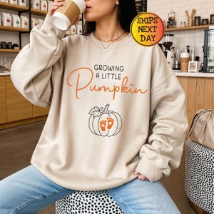 以下が含まれることがあります： ベージュのクルーネックトレーナー。胸元中央に黒とオレンジで「Growing a Little Pumpkin」の文字と、オレンジ色の赤ちゃんの足跡が描かれたカボチャの線画がプリントされています。長袖のプルオーバーです。
