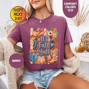 May include: A berry-colored short-sleeve t-shirt featuring a graphic print with the text 'It's Fall Y'all'. The design includes a cowboy hat, floral elements, a blue pumpkin, an orange pumpkin, and a pair of floral-patterned cowboy boots.