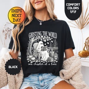 May include: A black t-shirt featuring a white graphic print of a ghost sitting in a chair under a tree reading a book. The text reads GHOSTING THE WORLD one chapter at a time. The design includes pumpkins, a lantern, and a stack of books.