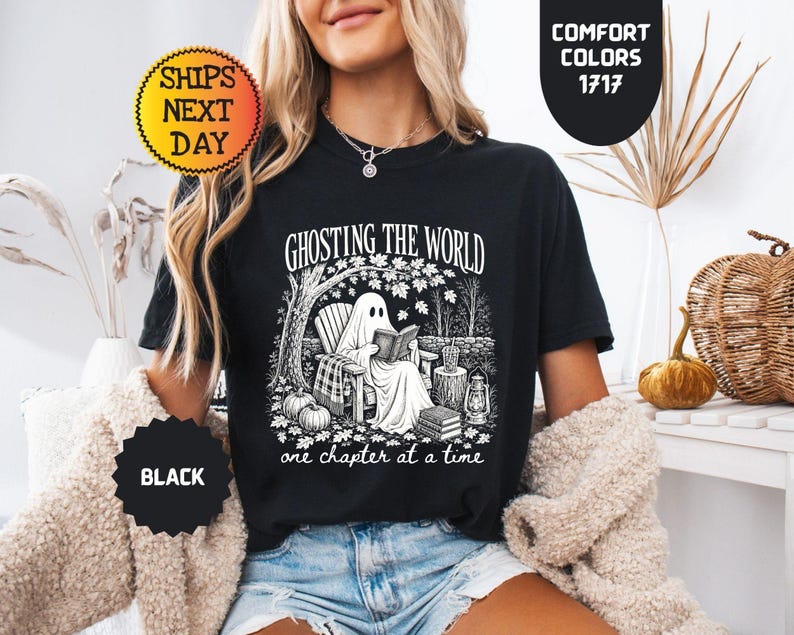 May include: A black t-shirt featuring a white graphic print of a ghost sitting in a chair under a tree reading a book. The text reads GHOSTING THE WORLD one chapter at a time. The design includes pumpkins, a lantern, and a stack of books.