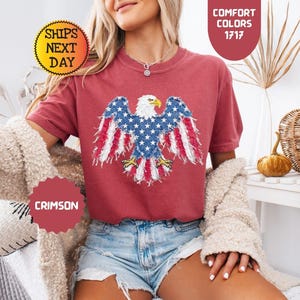 May include: Crimson-colored t-shirt featuring a patriotic design of an American bald eagle with red and white striped wings and a blue body with white stars. The shirt is part of the Comfort Colors collection.