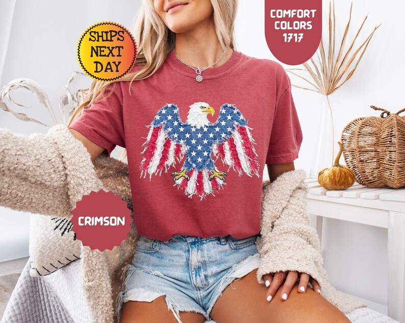 May include: Crimson-colored t-shirt featuring a patriotic design of an American bald eagle with red and white striped wings and a blue body with white stars. The shirt is part of the Comfort Colors collection.