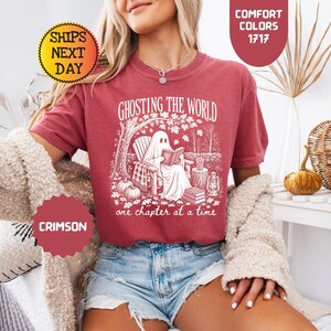 May include: A crimson short-sleeve t-shirt featuring a white graphic of a ghost sitting in a chair reading a book under a tree. The text reads GHOSTING THE WORLD one chapter at a time. The design includes pumpkins, a lantern, and fallen leaves.