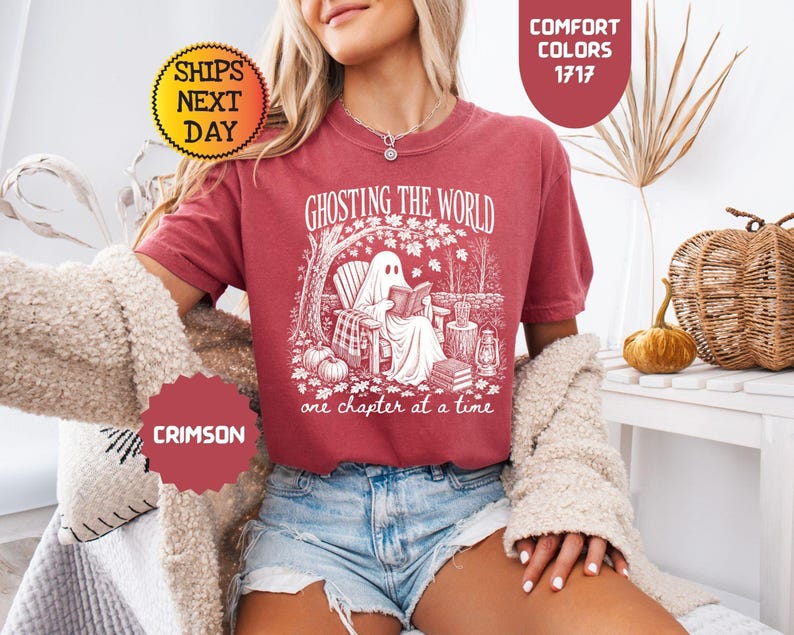 May include: A crimson short-sleeve t-shirt featuring a white graphic of a ghost sitting in a chair reading a book under a tree. The text reads GHOSTING THE WORLD one chapter at a time. The design includes pumpkins, a lantern, and fallen leaves.