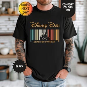 以下が含まれることがあります： 黒いTシャツで、金色のフォントで「Disney Dad」という文字が書かれ、その下にマルチカラーのストライプが入った垂直のバーコードデザインに統合された城のシルエットのグラフィックがあり、その下に「SCAN FOR PAYMENT」という文字が金色で印刷されています。