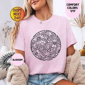 May include: A light pink t-shirt featuring a black and white floral volleyball design. The shirt has short sleeves and a crew neck. The text "COMFORT COLORS 1717" is in a pink oval. The text "SHIPS NEXT DAY" is in a yellow circle.