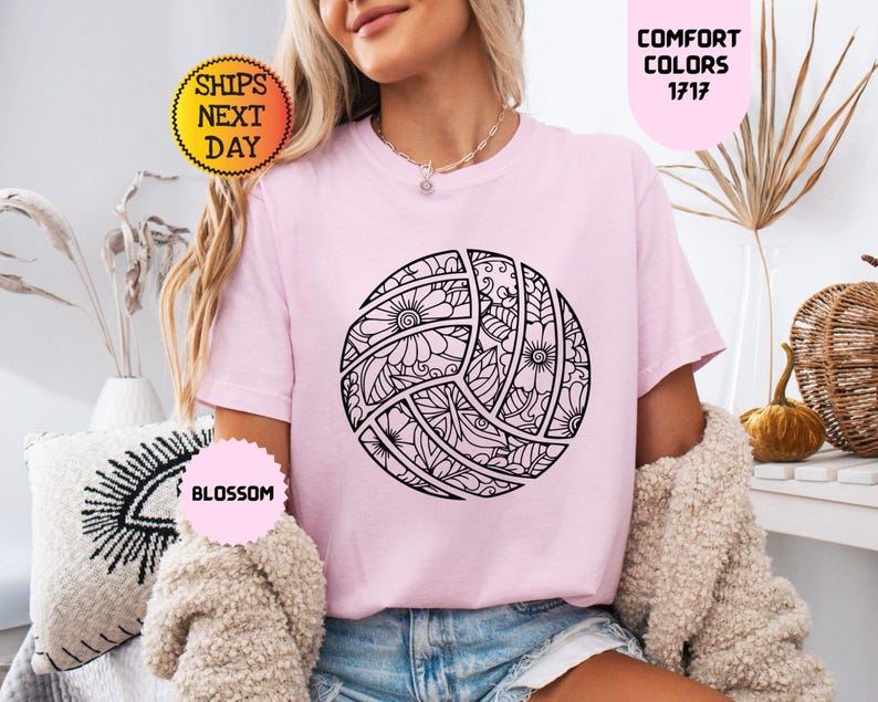 May include: A light pink t-shirt featuring a black and white floral volleyball design. The shirt has short sleeves and a crew neck. The text "COMFORT COLORS 1717" is in a pink oval. The text "SHIPS NEXT DAY" is in a yellow circle.