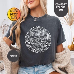 May include: A dark gray t-shirt featuring a white volleyball design with floral and geometric patterns. The shirt has the text "COMFORT COLORS 1717" and "SHIPS NEXT DAY". The shirt is labeled "PEPPER".