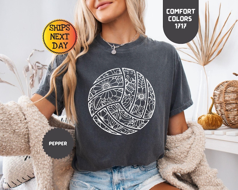May include: A dark gray t-shirt featuring a white volleyball design with floral and geometric patterns. The shirt has the text "COMFORT COLORS 1717" and "SHIPS NEXT DAY". The shirt is labeled "PEPPER".