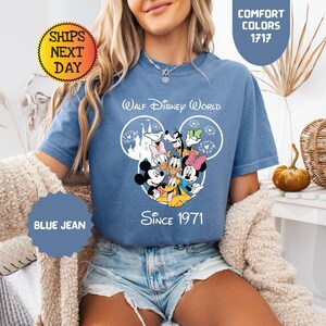 Pode incluir: Camiseta azul jeans com um gr&aacute;fico de personagens da Disney, incluindo Mickey e Minnie Mouse, Pato Donald e Pateta. A camisa tem o texto "Walt Disney World Since 1971". A camisa &eacute; uma cor de conforto.