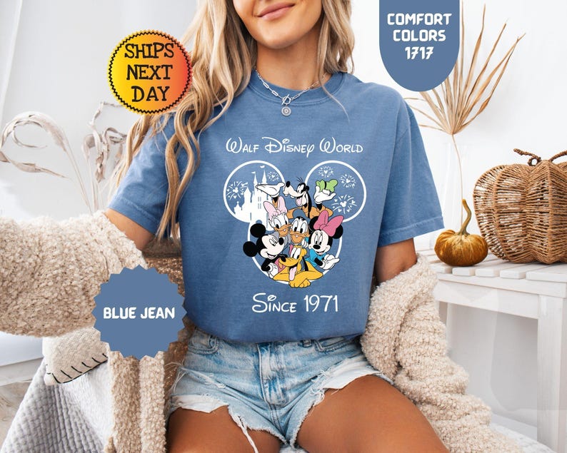 Pode incluir: Camiseta azul jeans com um gr&aacute;fico de personagens da Disney, incluindo Mickey e Minnie Mouse, Pato Donald e Pateta. A camisa tem o texto "Walt Disney World Since 1971". A camisa &eacute; uma cor de conforto.