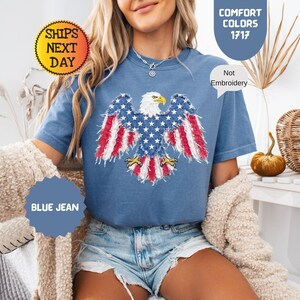 May include: Blue jean t-shirt featuring a distressed American flag eagle design. The eagle has a white head, yellow beak, and wings with red and white stripes and blue stars. The shirt is labeled "Comfort Colors 1717" and "Not Embroidery".