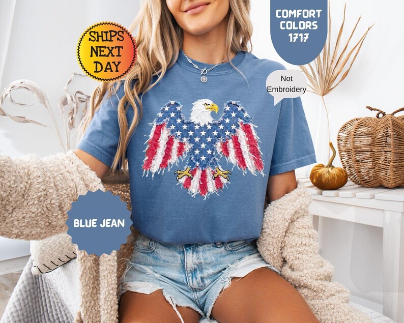 May include: Blue jean t-shirt featuring a distressed American flag eagle design. The eagle has a white head, yellow beak, and wings with red and white stripes and blue stars. The shirt is labeled "Comfort Colors 1717" and "Not Embroidery".