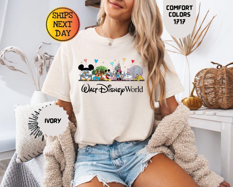 May include: Ivory-colored t-shirt featuring a colorful graphic of Disney characters and the words "Walt Disney World." The shirt has a crew neck and short sleeves. The image also includes the text "Comfort Colors 1717" and "Ships Next Day."