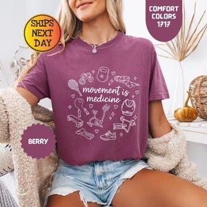 May include: Berry-colored t-shirt with the phrase "movement is medicine" surrounded by white line-art illustrations of fitness equipment. The shirt is paired with denim shorts. The image also includes text that says "Comfort Colors 1717" and "Ships Next Day".