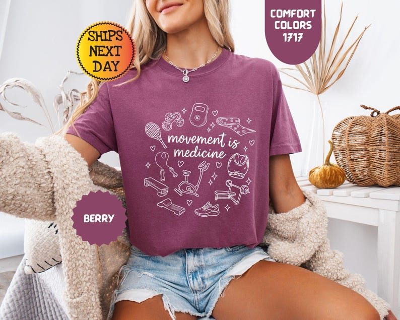 May include: Berry-colored t-shirt with the phrase "movement is medicine" surrounded by white line-art illustrations of fitness equipment. The shirt is paired with denim shorts. The image also includes text that says "Comfort Colors 1717" and "Ships Next Day".