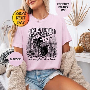 May include: A light pink t-shirt featuring a black graphic print of a ghost sitting in a chair under a tree reading a book. The text reads GHOSTING THE WORLD one chapter at a time. The shirt is labeled Comfort Colors 1717 and the color is Blossom.