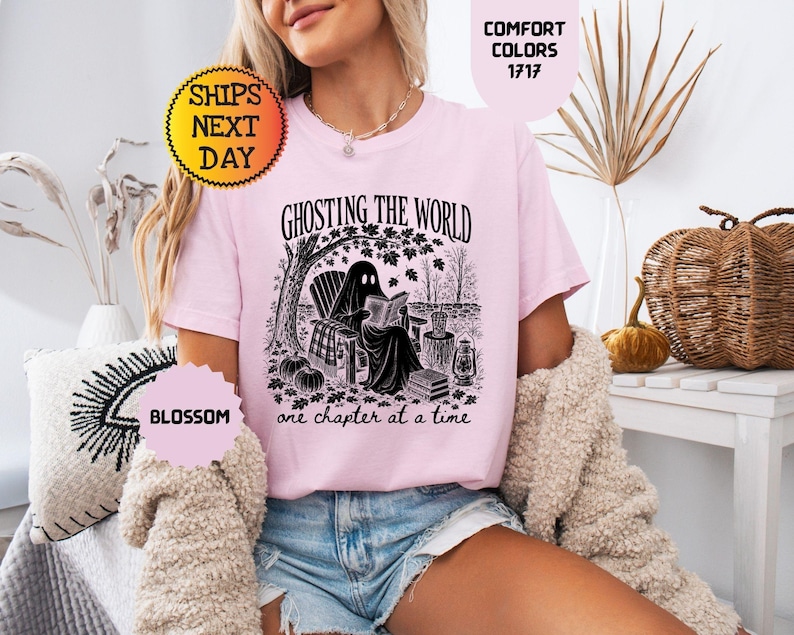 May include: A light pink t-shirt featuring a black graphic print of a ghost sitting in a chair under a tree reading a book. The text reads GHOSTING THE WORLD one chapter at a time. The shirt is labeled Comfort Colors 1717 and the color is Blossom.