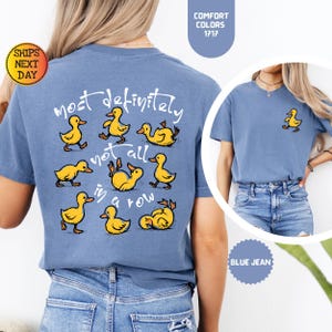 May include: Blue jean-colored t-shirt featuring a playful design of yellow cartoon ducks. The back of the shirt displays the text "most definitely not all in a row" above and around the ducks. A smaller duck graphic is on the front.