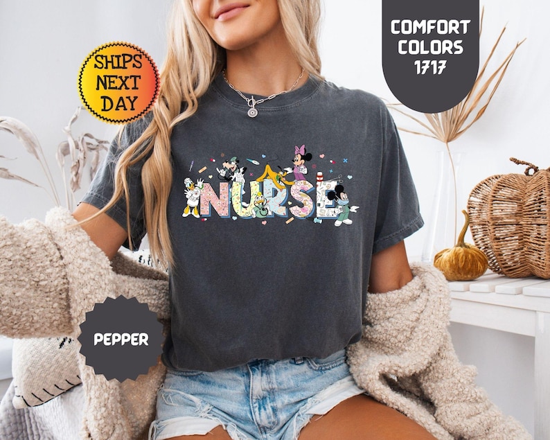 May include: A dark gray t-shirt with the word "NURSE" in colorful letters, featuring cartoon characters. The shirt has a round neck and short sleeves. The text "COMFORT COLORS 1717" is on the upper right.