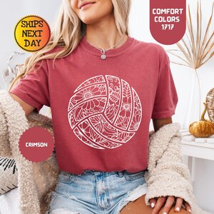 May include: Crimson-colored t-shirt featuring a white volleyball design with floral patterns. The shirt has short sleeves and is paired with denim shorts. The image also includes text that says "Comfort Colors 1717" and "Crimson".