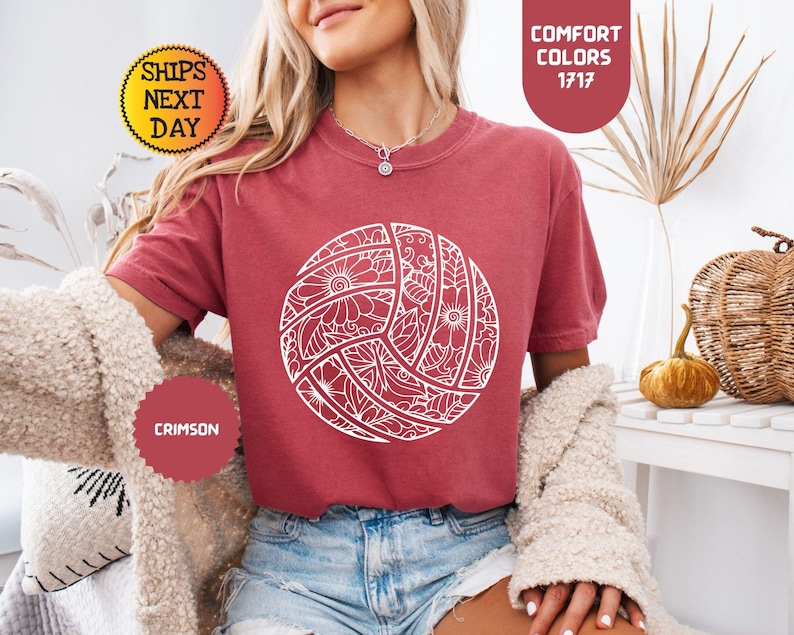 May include: Crimson-colored t-shirt featuring a white volleyball design with floral patterns. The shirt has short sleeves and is paired with denim shorts. The image also includes text that says "Comfort Colors 1717" and "Crimson".