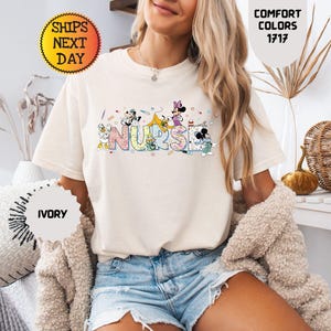 May include: Ivory-colored t-shirt with the word "NURSE" in colorful, cartoon-style letters. The letters are decorated with cartoon characters. The shirt is paired with denim shorts. The image also includes text that says "Comfort Colors 1717".