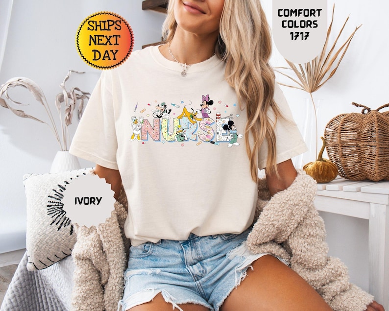 May include: Ivory-colored t-shirt with the word "NURSE" in colorful, cartoon-style letters. The letters are decorated with cartoon characters. The shirt is paired with denim shorts. The image also includes text that says "Comfort Colors 1717".