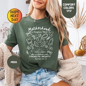 May include: A moss green t-shirt with the text "Motherhood is Kingdom Work" and other graphics in white. The shirt has short sleeves and is made of a soft material. The shirt is part of the "Comfort Colors" collection.