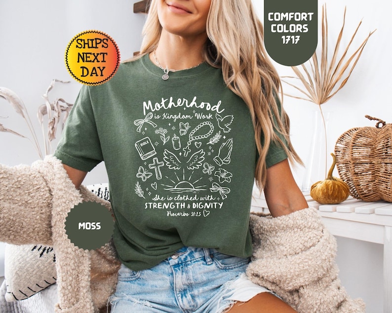 May include: A moss green t-shirt with the text "Motherhood is Kingdom Work" and other graphics in white. The shirt has short sleeves and is made of a soft material. The shirt is part of the "Comfort Colors" collection.