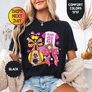 May include: A black t-shirt featuring a graphic design with a softball, a baseball jersey with the text NAME 21, a baseball bat, a glove, and pink and yellow checkered bows. The design includes stars and flowers in bright pink, yellow, and white.