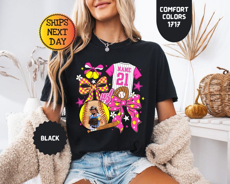 May include: A black t-shirt featuring a graphic design with a softball, a baseball jersey with the text NAME 21, a baseball bat, a glove, and pink and yellow checkered bows. The design includes stars and flowers in bright pink, yellow, and white.
