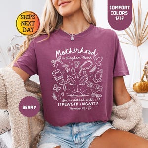 May include: Berry-colored t-shirt with the text "Motherhood is Kingdom Work" and other white graphics. The shirt also has the text "She is clothed with Strength & Dignity Proverbs 31:25". The shirt is part of the Comfort Colors collection.