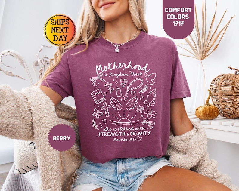 May include: Berry-colored t-shirt with the text "Motherhood is Kingdom Work" and other white graphics. The shirt also has the text "She is clothed with Strength & Dignity Proverbs 31:25". The shirt is part of the Comfort Colors collection.