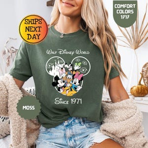 Pode incluir: T-shirt verde musgo com um design do Walt Disney World, apresentando Mickey Mouse, Minnie Mouse, Pato Donald e Pateta. A camisa inclui o texto "Walt Disney World Since 1971".