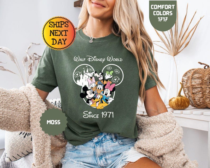 Pode incluir: T-shirt verde musgo com um design do Walt Disney World, apresentando Mickey Mouse, Minnie Mouse, Pato Donald e Pateta. A camisa inclui o texto "Walt Disney World Since 1971".